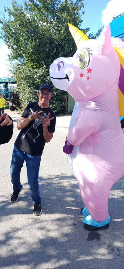 Ein Mann in schwarzem T-Shirt und Baseballmütze posiert mit einer Person in einem bunten, aufblasbaren Einhornkostüm im Freien an einem sonnigen Tag.