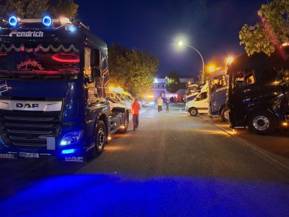 In einer beleuchteten Nachtstraße stehen links und rechts mehrere große, beleuchtete Lkw und Kleintransporter, während einige Menschen zu Fuß zwischen den Fahrzeugen gehen.