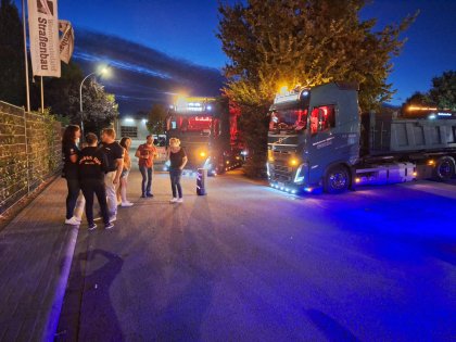 Sechs Personen stehen am Straßenrand bei Dunkelheit auf einer Straße mit zwei beleuchteten Lkw, während im Hintergrund Fahnen mit der Aufschrift „Westministerium Straßenbau“ wehen.