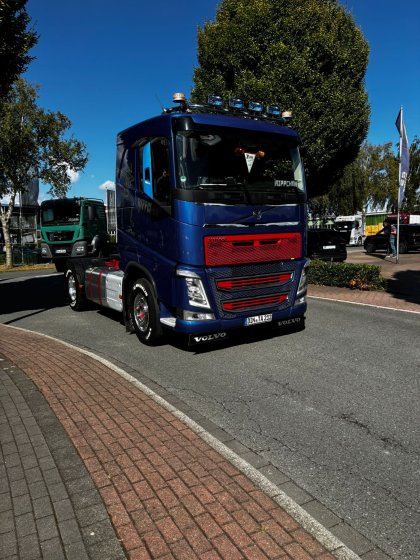 Blau-roter Volvo-Sattelzug mit rotem Kühlergrill und Kennzeichen DIN TA 212 fährt auf einer Straße mit rotem Gehweg bei sonnigem Wetter.